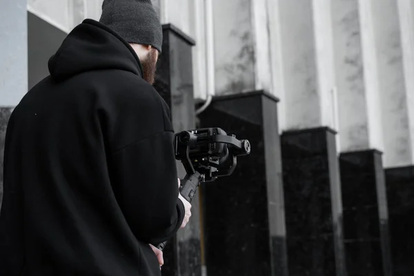 Vollbärtiger professioneller Videofilmer in schwarzem Kapuzenpullover, der eine professionelle Kamera auf einem 3-Achsen-Gimbal-Stabilisator hält. Filmemacher, der ein großartiges Video mit einer professionellen Kinokamera macht. Kameramann. — Stockfoto