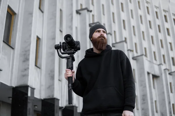 Vollbärtiger professioneller Videofilmer in schwarzem Kapuzenpullover, der eine professionelle Kamera auf einem 3-Achsen-Gimbal-Stabilisator hält. Filmemacher, der ein großartiges Video mit einer professionellen Kinokamera macht. Kameramann. — Stockfoto