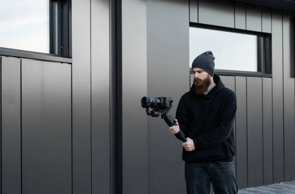 Vídeo profissional barbudo com capuz preto segurando câmera profissional no estabilizador de 3 eixos gimbal. Filmmaker fazendo um grande vídeo com uma câmera de cinema profissional. Fotografo . — Fotografia de Stock