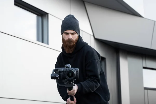 Brodaty Profesjonalny fotograf w czarnej bluzie z kapturem trzymający profesjonalny aparat na 3-osiowym stabilizatorze gimbalu. Filmowiec robi świetny film z profesjonalną kamerą kinową. Operator filmowy. — Zdjęcie stockowe