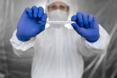 Beyaz koruyucu giysili doktor, tıbbi maske ve lastik eldivenler koronavirüs salgınında termometre tutuyor. Salgın, Coronavirus covid 19 salgını. Sıcaklık ölçülüyor ve gösteriliyor.