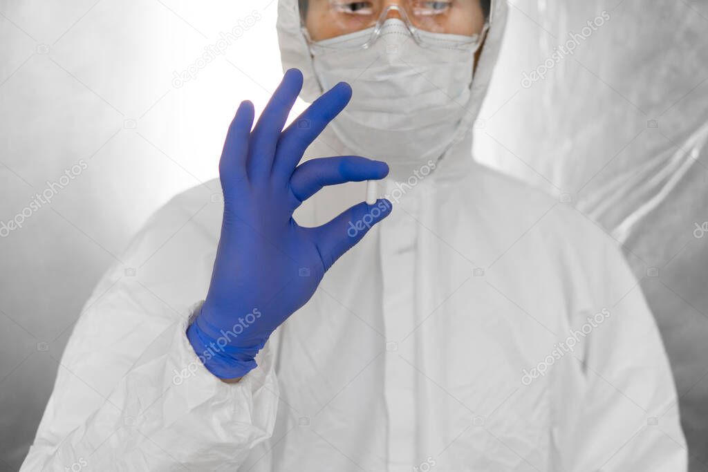Doctor in white protective suit, medical mask and rubber gloves is holding a white pill as a medications from coronavirus pandemic threat. Epidemic, pandemic of coronavirus covid 19.