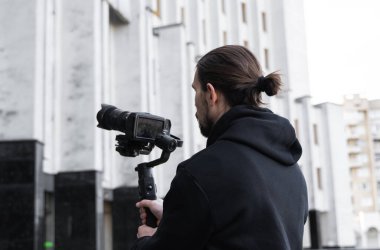 3 eksenli titreşimli dengeleyicide profesyonel kamera tutan genç profesyonel kameraman. Pro ekipmanlar titremeden yüksek kaliteli video çekmeye yardımcı olur. Siyah kapüşonlu bir kameraman video çekiyor..