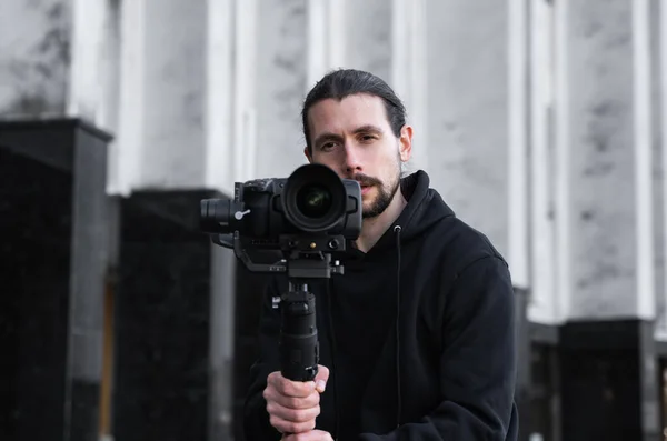 Fiatal profi videós professzionális kamerát tart 3 tengelyes gimbal stabilizátoron. Pro berendezések segít, hogy kiváló minőségű videót rázás nélkül. Fekete kapucnis operatőr videókat készít. — Stock Fotó