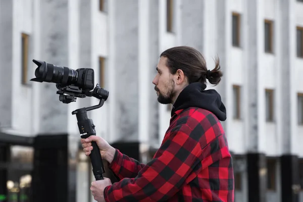 Νέοι επαγγελματίες videographer κρατώντας επαγγελματική φωτογραφική μηχανή σε 3-άξονα gimbal σταθεροποιητή. Pro εξοπλισμός βοηθά να κάνει υψηλής ποιότητας βίντεο χωρίς ανακίνηση. Καμεραμάν φορώντας κόκκινο πουκάμισο κάνοντας βίντεο. — Φωτογραφία Αρχείου