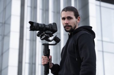 3 eksenli titreşimli dengeleyicide profesyonel kamera tutan genç profesyonel kameraman. Pro ekipmanlar titremeden yüksek kaliteli video çekmeye yardımcı olur. Beyaz kapüşonlu bir kameraman video çekiyor..
