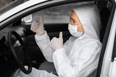 Koruyucu giysili, tıbbi maskeli ve virüs ve bakterilerden korunmak için lastik eldivenli bir adam bir arabada oturuyor ve baş parmağını kaldırıyor. Karantina, dünya salgını, covid 19, koronavirüs, enfeksiyon..