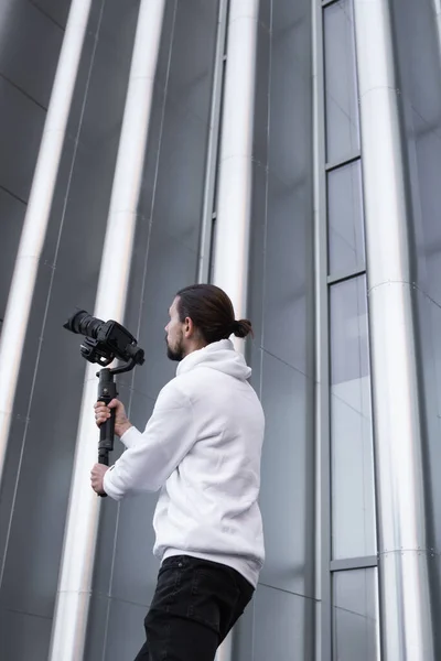 Ung professionell videograf som håller professionell kamera på 3-axlig gimbal stabilisator. Pro-utrustning hjälper till att göra högkvalitativ video utan att skaka. Kameraman bär vit luvtröja gör en video. — Stockfoto