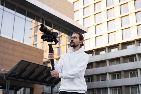 Giovane videografo professionale con fotocamera professionale su stabilizzatore gimbale a 3 assi. L'attrezzatura Pro aiuta a fare video di alta qualità senza scuotere. Cameraman indossa felpa bianca facendo un video . — Foto Stock