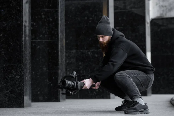 Vollbärtiger professioneller Videofilmer in schwarzem Kapuzenpullover, der eine professionelle Kamera auf einem 3-Achsen-Gimbal-Stabilisator hält. Filmemacher, der ein großartiges Video mit einer professionellen Kinokamera macht. Kameramann. — Stockfoto