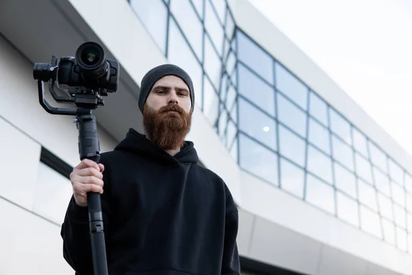 Γενειάδα επαγγελματική videographer σε μαύρο hoodie κρατώντας επαγγελματική κάμερα σε 3-άξονα gimbal σταθεροποιητή. Κινηματογραφιστής κάνει ένα μεγάλο βίντεο με μια επαγγελματική κάμερα κινηματογράφου. Κινηματογράφος. — Φωτογραφία Αρχείου