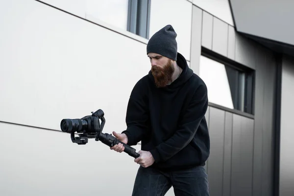 Vídeo profissional barbudo com capuz preto segurando câmera profissional no estabilizador de 3 eixos gimbal. Filmmaker fazendo um grande vídeo com uma câmera de cinema profissional. Fotografo . — Fotografia de Stock