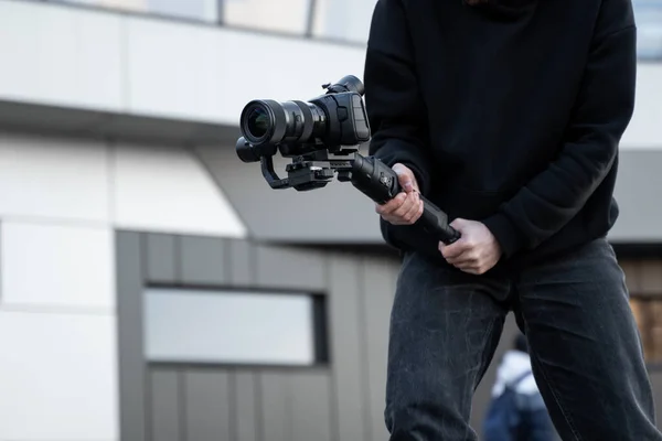 Профессиональный видеооператор в черной толстовке держит профессиональную камеру на 3-осном стабилизаторе. Режиссёр снимает отличное видео с профессиональной кинокамерой. Кинооператор . — стоковое фото
