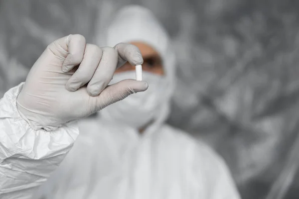 Doutor em terno de proteção branco, máscara médica e luvas de borracha está segurando uma pílula branca como medicamentos contra a ameaça de pandemia de coronavírus. Epidemia, pandemia de coronavírus covid 19 . — Fotografia de Stock