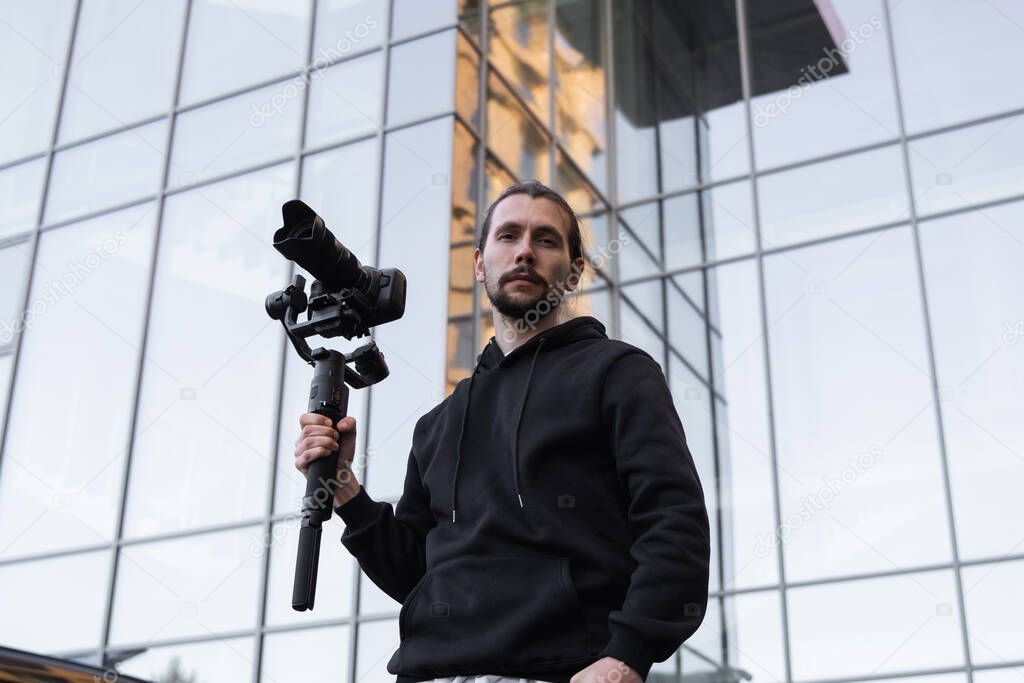 Young Professional videographer holding professional camera on 3-axis gimbal stabilizer. Pro equipment helps to make high quality video without shaking. Cameraman wearing white hoodie making a videos.