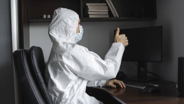 Tıbbi maskeli ve koruyucu elbiseli bir adam, eldivenler evde oturuyor ve karantina sırasında bilgisayarda çalışıyor ve iş ortağına ya da arkadaşlarına baş parmağını gösteriyor. Bir salgın covid içinde uzaktan çalışma.