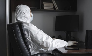 Tıbbi maskeli ve koruyucu giysili bir adam, lastik eldivenler evde oturuyor ve karantina sırasında masada bilgisayarla çalışıyor. Tasarımcı, sanatçı, mimar, iş adamı salgın bir covid 'de uzaktan çalışıyor..