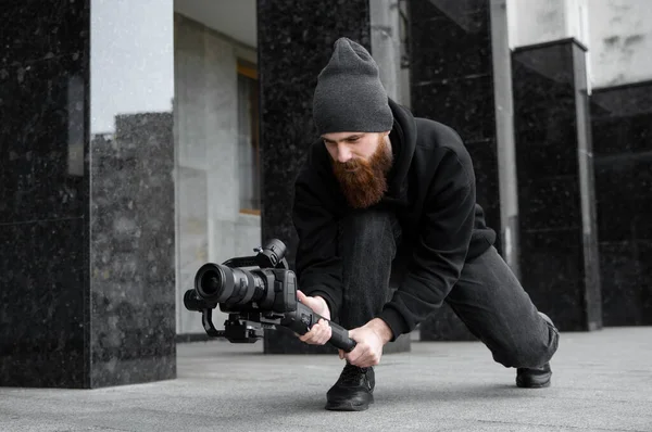 Vollbärtiger professioneller Videofilmer in schwarzem Kapuzenpullover, der eine professionelle Kamera auf einem 3-Achsen-Gimbal-Stabilisator hält. Filmemacher, der ein großartiges Video mit einer professionellen Kinokamera macht. Kameramann. — Stockfoto