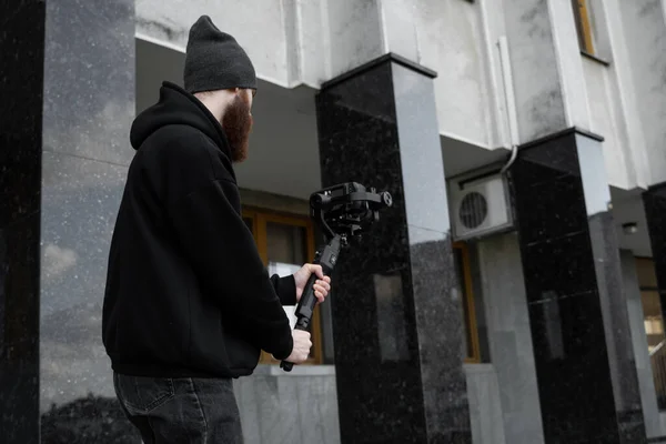 Bearded Professional videographer in black hoodie holding professional camera on 3-axis gimbal stabilizer. Filmmaker making a great video with a professional cinema camera. Cinematographer. — Stock Photo, Image