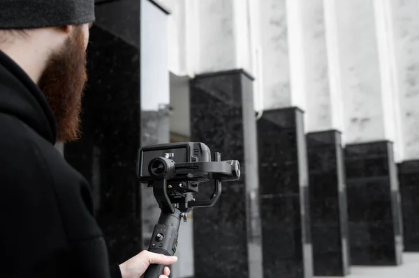 Vidéographe professionnel barbu en sweat à capuche noir tenant une caméra professionnelle sur stabilisateur de cardan 3 axes. Cinéaste faire une grande vidéo avec une caméra de cinéma professionnel. Directeur de la photographie . — Photo