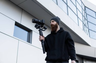 Siyah kapüşonlu sakallı profesyonel kameraman elinde 3 eksenli titreşim dengeleyicisi var. Film yapımcısı profesyonel bir sinema kamerasıyla harika bir video çekiyor. Görüntü Yönetmeni.