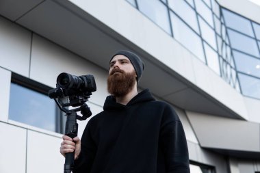 Siyah kapüşonlu sakallı profesyonel kameraman elinde 3 eksenli titreşim dengeleyicisi var. Film yapımcısı profesyonel bir sinema kamerasıyla harika bir video çekiyor. Görüntü Yönetmeni.