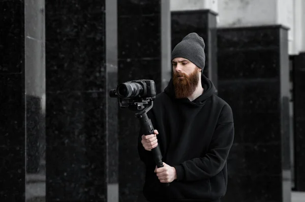 Vidéographe professionnel barbu en sweat à capuche noir tenant une caméra professionnelle sur stabilisateur de cardan 3 axes. Cinéaste faire une grande vidéo avec une caméra de cinéma professionnel. Directeur de la photographie . — Photo