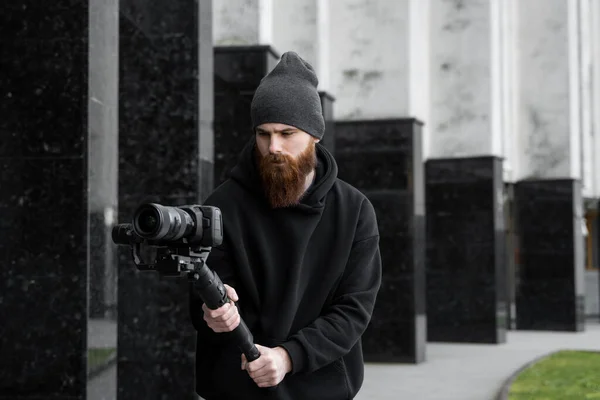 Γενειάδα επαγγελματική videographer σε μαύρο hoodie κρατώντας επαγγελματική κάμερα σε 3-άξονα gimbal σταθεροποιητή. Κινηματογραφιστής κάνει ένα μεγάλο βίντεο με μια επαγγελματική κάμερα κινηματογράφου. Κινηματογράφος. — Φωτογραφία Αρχείου