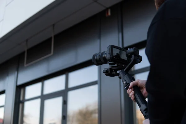 Vídeo profissional com capuz preto segurando câmera profissional no estabilizador de 3 eixos gimbal. Filmmaker fazendo um grande vídeo com uma câmera de cinema profissional. Fotografo . — Fotografia de Stock