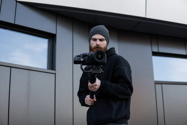 Videografo professionale barbuto con cappuccio nero con fotocamera professionale su stabilizzatore gimbale a 3 assi. Filmmaker fare un grande video con una macchina fotografica cinematografica professionale. Cinematografo . — Foto Stock