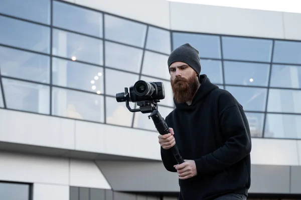 Bearded Professional videographer in black hoodie holding professional camera on 3-axis gimbal stabilizer. Filmmaker making a great video with a professional cinema camera. Cinematographer.