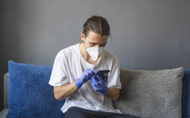 Tıbbi maskeli ve lastik eldivenli bir adam evde oturuyor ve karantina sırasında koltukta telefon kullanıyor. Tasarımcı, sanatçı, mimar, salgın bir covid 'in uzaktan kumandalı işadamı. Coronavirüs.