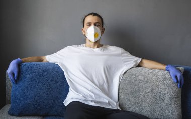 Beyaz gömlekli, tıbbi maskeli ve lastik eldivenli bir adam kanepede oturuyor ve salgın koronavirüsünü izlerken kendini harika hissediyor. Salgın, Coronavirus covid 19 salgını..