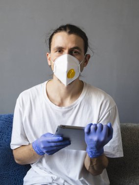 Beyaz gömlekli, tıbbi maskeli ve lastik eldivenli bir adam evde oturuyor ve karantina sırasında kanepede tabletle çalışıyor. İnsan, tasarımcı, sanatçı, mimar, iş adamı salgın bir covid 'de uzaktan çalışıyor.