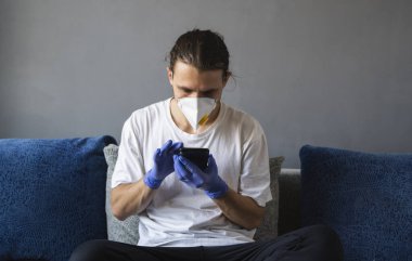 Tıbbi maskeli ve lastik eldivenli bir adam evde oturuyor ve karantina sırasında koltukta telefon kullanıyor. Tasarımcı, sanatçı, mimar, salgın bir covid 'in uzaktan kumandalı işadamı. Coronavirüs.