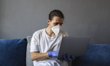 Tıbbi maskeli ve lastik eldivenli bir adam evde oturuyor ve karantina sırasında kanepede dizüstü bilgisayarla çalışıyor. Tasarımcı, sanatçı, mimar, salgın bir covid 'in uzaktan kumandalı işadamı. Coronavirüs.