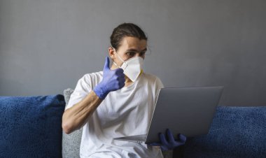 Tıbbi maskeli ve lastik eldivenli bir adam evde oturuyor ve karantina sırasında kanepede dizüstü bilgisayarla çalışıyor. Tasarımcı, sanatçı, mimar, salgın bir covid 'in uzaktan kumandalı işadamı. Coronavirüs.