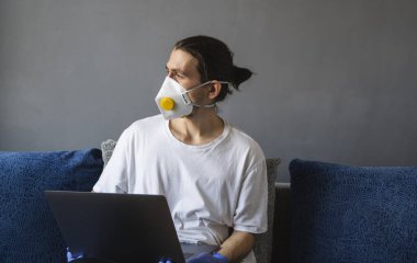Tıbbi maskeli ve lastik eldivenli bir adam evde oturuyor ve karantina sırasında kanepede dizüstü bilgisayarla çalışıyor. Tasarımcı, sanatçı, mimar, salgın bir covid 'in uzaktan kumandalı işadamı. Coronavirüs.