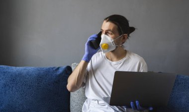 Tıbbi maskeli ve lastik eldivenli bir adam evde oturuyor ve karantina sırasında telefonla biriyle konuşuyor..