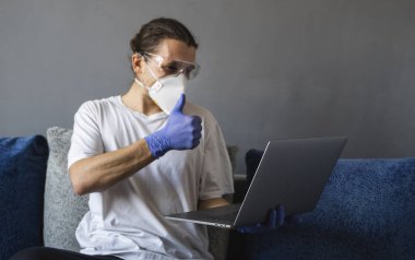 Tıbbi maskeli ve lastik eldivenli bir adam evde oturuyor ve karantina sırasında kanepede dizüstü bilgisayarla çalışıyor. Tasarımcı, sanatçı, mimar, salgın bir covid 'in uzaktan kumandalı işadamı. Coronavirüs.