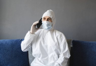Koruyucu takım elbiseli, tıbbi maskeli ve lastik eldivenli bir adam evde oturuyor ve karantina sırasında bir kanepede arkadaşlarıyla veya iş arkadaşlarıyla konuşuyor. Salgın bir covid 'de uzaktan çalışan bir adam..