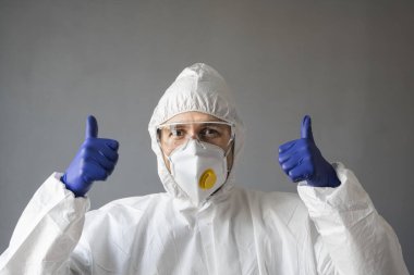 Beyaz koruyucu giysili, tıbbi maskeli ve lastik eldivenli doktor baş parmağını kaldırıyor. Koronavirüs salgını tehdidi altındayken insanlara yardım etmek. Salgın, Coronavirus covid 19 salgını..