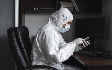Koruyucu giysili, tıbbi maskeli ve lastik eldivenli bir adam evde oturuyor ve karantina sırasında arkadaşlarıyla veya iş ortaklarıyla sohbet etmek için telefon kullanıyor. Salgın bir covid 'de uzaktan çalışan bir adam.