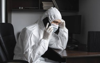 Koruyucu takım elbiseli, tıbbi maskeli ve lastik eldivenli bir adam evde oturuyor ve karantina sırasında arkadaşlarıyla veya iş ortağıyla telefonda konuşuyor. Uzaktan kumandalı bir adam salgın hastalığın pençesinde. Coronavirüs.