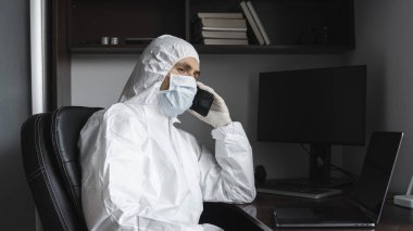 Koruyucu takım elbiseli, tıbbi maskeli ve lastik eldivenli bir adam evde oturuyor ve karantina sırasında arkadaşlarıyla veya iş ortağıyla telefonda konuşuyor. Uzaktan kumandalı bir adam salgın hastalığın pençesinde. Coronavirüs.