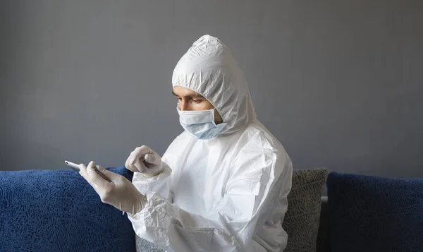 Man in protective suit, medical mask and rubber gloves sits at home and works with tablet on a sofa during quarantine. Designer, artist, architect, businessman at remote work in a pandemic covid.