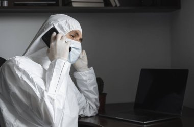 Koruyucu takım elbiseli, tıbbi maskeli ve lastik eldivenli bir adam evde oturuyor ve karantina sırasında arkadaşlarıyla veya iş ortağıyla telefonda konuşuyor. Uzaktan kumandalı bir adam salgın hastalığın pençesinde. Coronavirüs.