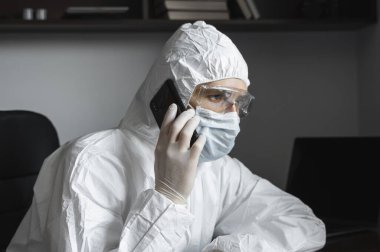 Koruyucu takım elbiseli, tıbbi maskeli ve lastik eldivenli bir adam evde oturuyor ve karantina sırasında arkadaşlarıyla veya iş ortağıyla telefonda konuşuyor. Uzaktan kumandalı bir adam salgın hastalığın pençesinde. Coronavirüs.