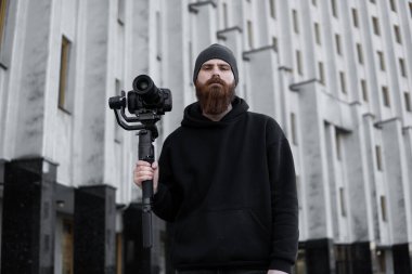 Siyah kapüşonlu sakallı profesyonel kameraman elinde 3 eksenli titreşim dengeleyicisi var. Film yapımcısı profesyonel bir sinema kamerasıyla harika bir video çekiyor. Görüntü Yönetmeni.