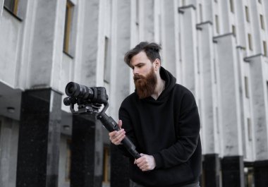 Siyah kapüşonlu sakallı profesyonel kameraman elinde 3 eksenli titreşim dengeleyicisi var. Film yapımcısı profesyonel bir sinema kamerasıyla harika bir video çekiyor. Görüntü Yönetmeni.